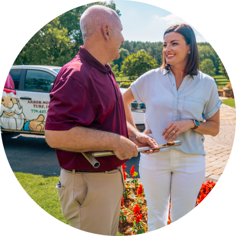 A lawn care professional holding a lawn tool, speaking with a smiling homeowner in front of a service truck and landscaped garden